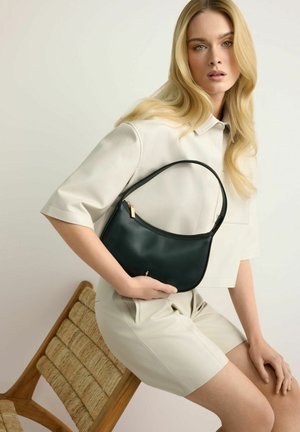 Blondynka w beżowym stroju siedząca na plecionym krześle, trzymająca czarną skórzaną torebkę z złotym zamkiem błyskawicznym i małym złotym detalem.