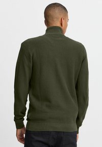 Grüner gestrickter Pullover mit hohem Kragen, mit strukturiertem Stoff, gerippten Bündchen und Saum. Sichtbares Logo auf der unteren Seite.