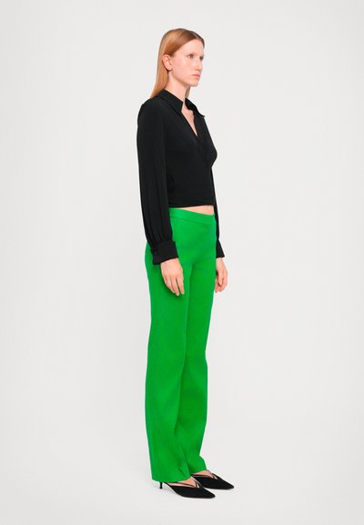 Blouse noire à manches longues avec des poignets boutonnés, associée à un pantalon droit vert vif et des escarpins noirs à bout pointu.