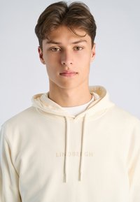 Jonge man met kort bruin haar, gekleed in een crèmekleurige hoodie met trekkoorden en een subtiele logotekst op de borst, neutrale achtergrond.