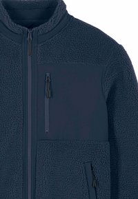 Veste polaire bleu foncé avec un col montant, un tissu texturé et deux poches zippées. Comprend un panneau lisse au niveau de la poitrine et une fermeture éclair.