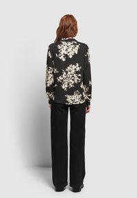 Schwarze Bluse mit weißem Blumenmuster, langen Ärmeln, Kragen und lockerer Passform. Kombiniert mit schwarzen weiten Hosen.