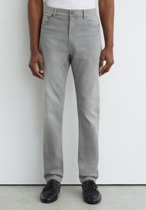 Homme portant un jean droit gris clair et des chaussures noires à lacets, debout contre un mur blanc texturé.