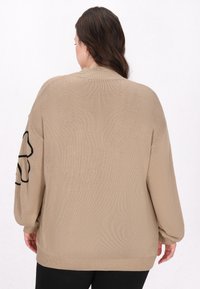 Gilet en tricot beige avec une coupe décontractée, des manches ballons et un motif floral noir sur la manche gauche. Tissu texturé avec un ourlet côtelé.