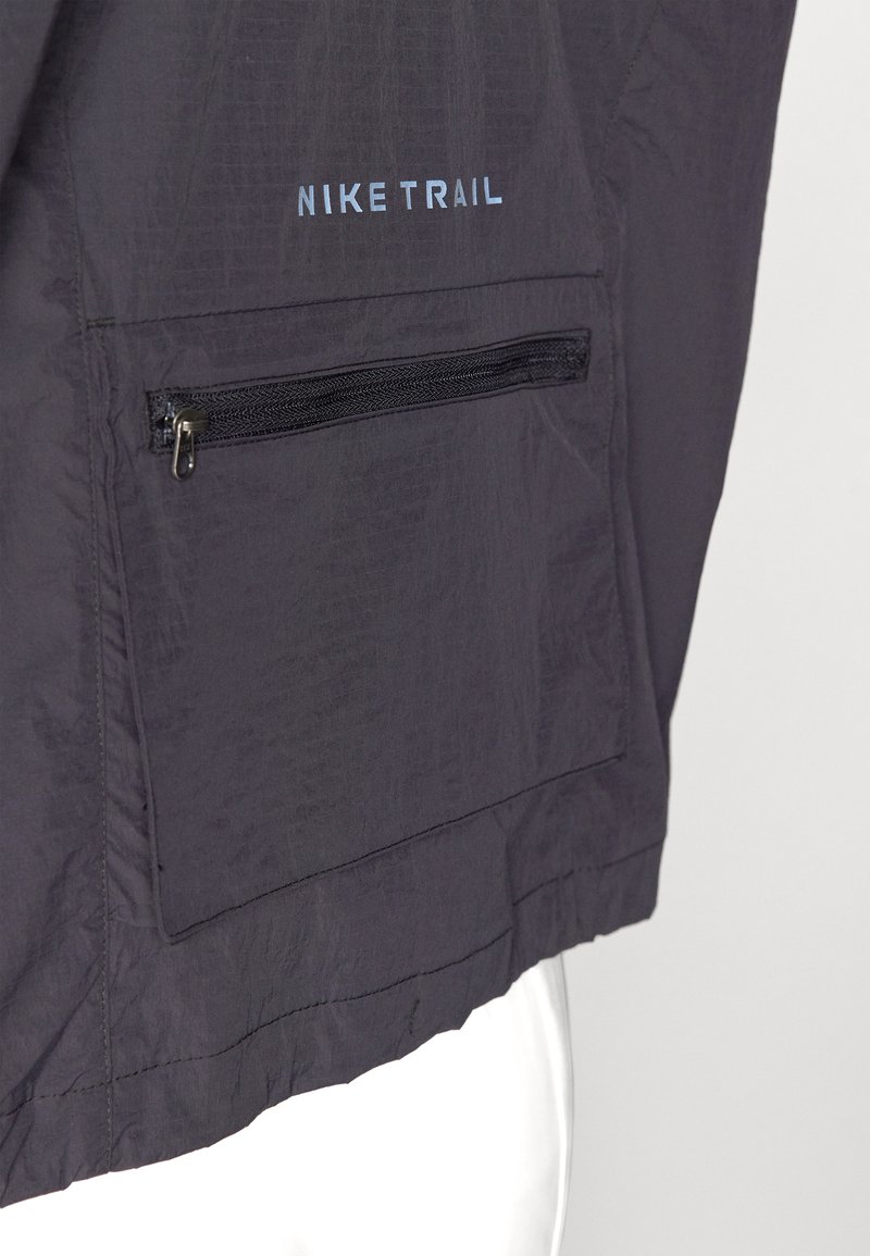 Nike Trail-Jacke aus dunkelgrauem Stoff, mit einer Reißverschlusstasche mit schwarzem Besatz und einem geprägten Logo in Hellblau über der Tasche.