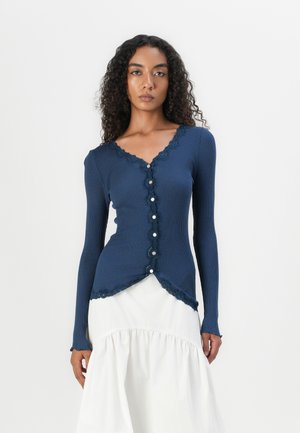 Marineblauwe ribgebreide cardigan met lange mouwen, kantafwerking en knoopsluiting. Heeft een gebogen zoom en past nauwsluitend om het lichaam. Draag het over een witte rok.