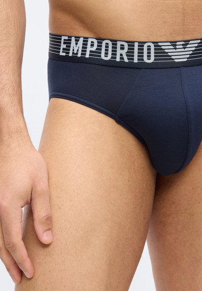 Άνδρας με σκούρα μπλε εσώρουχα με φαρδιά μαύρη ζώνη μέσης που φέρει το κείμενο "EMPORIO" και λογότυπο, στέκεται με ορατό το αριστερό χέρι.