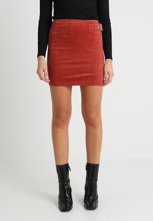 Woman wearing a rust-colored corduroy mini skirt with side buckle and black long-sleeve top paired with black ankle boots.
