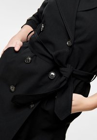 Abrigo negro tipo trench con diseño de doble botonadura, grandes botones y cinturón atado en la cintura. Tela suave con un corte entallado.