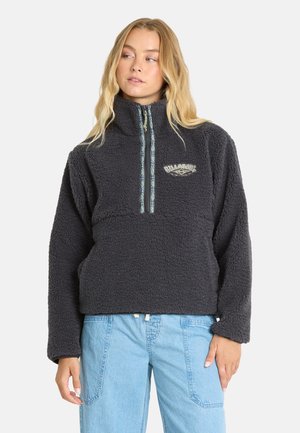 Vrouw draagt een donkergrijze fleece pullover met een half-rits met patroon en lichtblauwe spijkerbroek, kijkt naar links tegen een effen achtergrond.