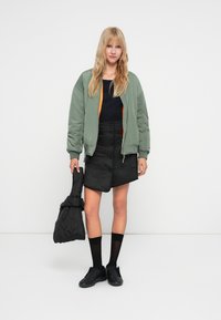 Veste bomber vert olive avec doublure orange, haut noir, jupe noire texturée avec des boutons, baskets noires et un sac assorti.