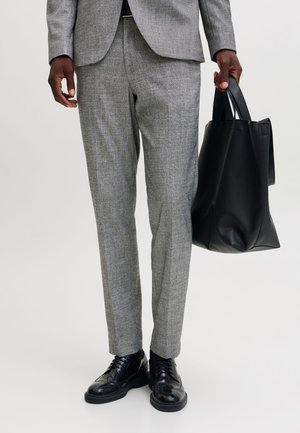 Une personne en costume gris et chaussures de ville noires tient un grand sac cabas en cuir noir sur un fond uni.