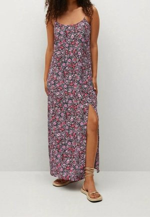 Femme portant une robe maxi fleurie sans manches avec une fente latérale et des sandales plates beiges à lacets, debout devant un fond clair uni.