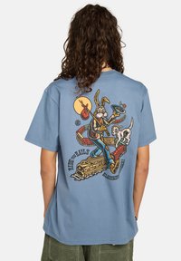 Person med langt krøllet hår iført en blå T-shirt med en tegneseriekanin, der holder en hat med en blomst, går på skinner med en hund.