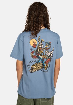 Persona con cabello largo y rizado lleva una camiseta azul con un dibujo de un conejo de dibujos animados sosteniendo un sombrero con una flor, caminando por las vías del tren con un perro.