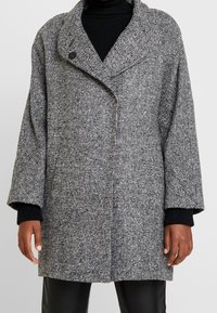 Grijze en zwarte houndstooth-gepatroneerde jas met een brede kraag, oversized silhouet en een enkele knoopsluiting. Beschikt over textuurstof en losse mouwen.
