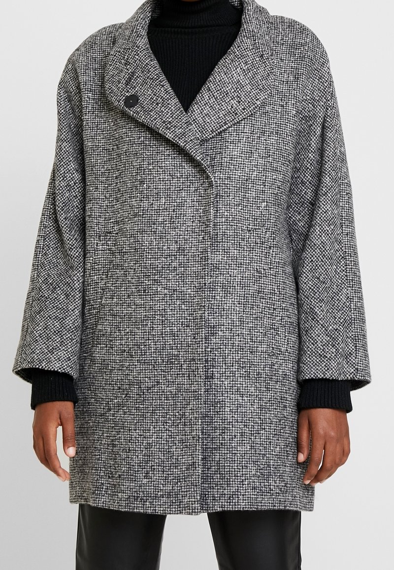 Grijze en zwarte houndstooth-gepatroneerde jas met een brede kraag, oversized silhouet en een enkele knoopsluiting. Beschikt over textuurstof en losse mouwen.