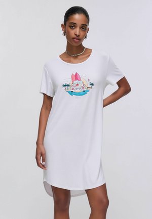 Donna con abito bianco con grafica colorata di tavola da surf, macchina, palme e faro sul davanti su sfondo semplice.