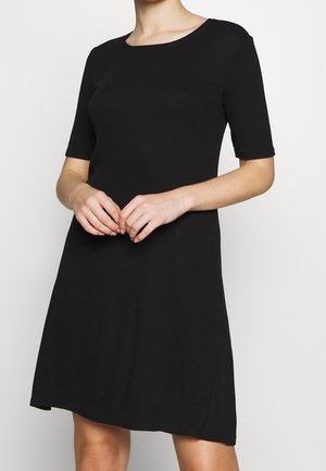 Femme portant une robe noire à manches courtes, longueur genoux, debout, les mains doucement jointes à la taille, sur un fond blanc uni.