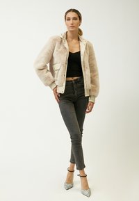 Beige faux fur bomberjakke med en cremefarget foring, trykknaplukninger, ribbede manchetter, parret med sorte skinny jeans og sølvhæle.