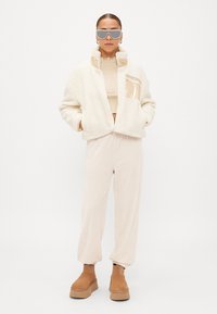 Casaco creme felpudo com um bolso brilhante, combinado com uma blusa bege e calças de jogger a condizer. Botas de plataforma castanhas completam o visual.