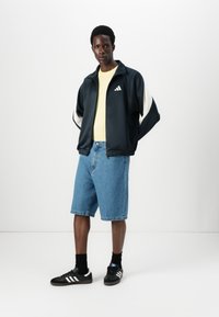 Casaco de fato de treino azul-marinho com detalhes brancos, camisa amarela, calções de ganga e ténis pretos com riscas brancas; tecido suave e corte descontraído.