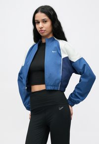 Veste bleue raccourcie avec accents blancs et logo Nike, dotée de poignets élastiques et d'une fermeture éclair, portée sur un haut noir et un legging.