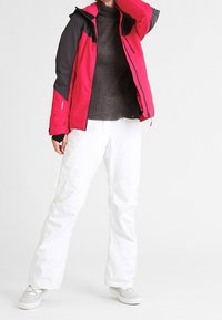 Giacca da sci rossa e nera con cappuccio, maglione scuro testurizzato e pantaloni da sci bianchi. Design elegante con colori a contrasto e dettagli riflettenti.