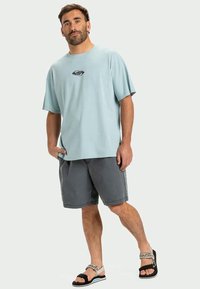 Hele sinine puuvillane T-särk, mille keskosas on väike graafika, koos tumedate hallide lühikeste pükste ja mustade sandaalidega, millel on mustriline rihm.