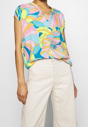 Kvinde iført en kortærmet bluse med et farverigt abstrakt blomsterprint og offwhite bukser med høj talje mod en ensfarvet baggrund.