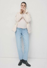 Kobieta ubrana w kremową polarową kurtkę, jasnoróżowy golf, jasnoniebieskie jeansy i czarne botki, stojąca na tle białej ściany.