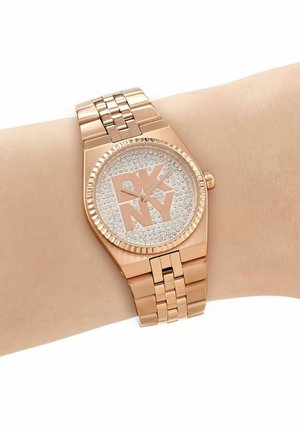 Orologio da polso in metallo color oro rosa con cinturino a maglie, quadrante tempestato di cristalli e grande logo DKNY al centro sul polso di una persona.