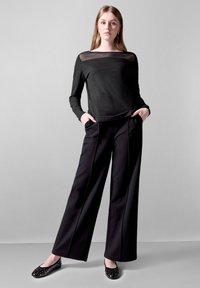 Top nero a maniche lunghe con scollatura trasparente, abbinato a pantaloni neri a gamba larga. Le calzature includono ballerine nere con dettagli borchiati.