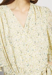 Světlě žluté šaty s květinovým vzorem, s výstřihem do V, krátkými rukávy a nařaseným detailem v pase.