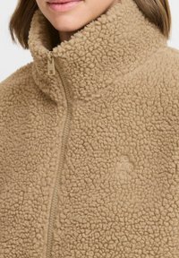 Veste en polaire beige avec un col montant et une fermeture éclair. Elle présente une texture douce et un branding subtil, mettant en avant un design moelleux et confortable.