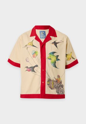 Homme portant une chemise beige et rouge à manches courtes avec un motif d’oiseaux et de paniers, un short blanc avec un texte manuscrit bleu, des chaussettes blanches et des chaussures noires.