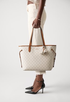 Una persona è in piedi con pantaloni dai toni neutri, tiene una borsa tote elegante con un design a motivi e frange, indossando tacchi alti neri.
