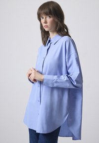 Cămașă albastră deschis, cu mânecă lungă, cu închidere prin butoni, guler clasic, hem posterior prelungit și croi lejer, realizată dintr-un material neted.