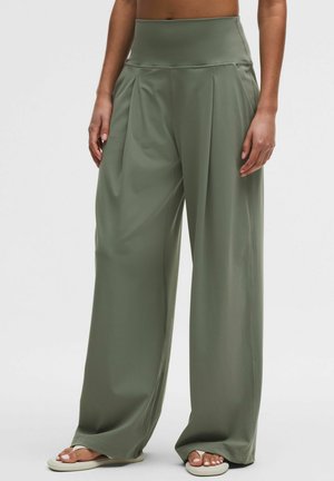 Kvinde iført højtaljede, løstsiddende olivengrønne brede bukser og hvide sandaler, stående mod en ensfarvet lys baggrund.