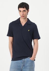Mörkblå polo med krage, korta ärmar och en gul broderad logotyp på vänster bröst; bärs med ljusblå jeans.