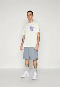 Bela bomba majica iz bombaža z vijoličnim grafičnim žepom, v kombinaciji s sivimi šortami. Opremljena z belimi supergami in sivimi nogavicami, s tetovažami na rokah.