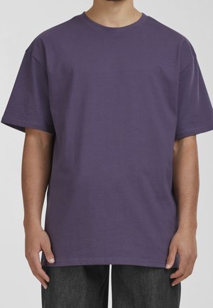 Lila Baumwoll-T-Shirt, kurze Ärmel, Rundhalsausschnitt, lockere Passform. Einfaches Design ohne Muster oder Grafiken, gehalten von einer Person, die dunkle Jeans trägt.