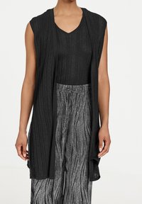 Cardigan nera senza maniche con texture a coste e scollo aperto, indossato sopra un top nero a scollo a V e pantaloni a fantasia bianco e nero.