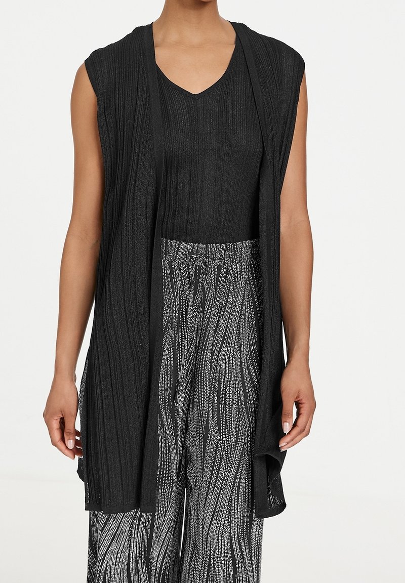 Cardigan nera senza maniche con texture a coste e scollo aperto, indossato sopra un top nero a scollo a V e pantaloni a fantasia bianco e nero.