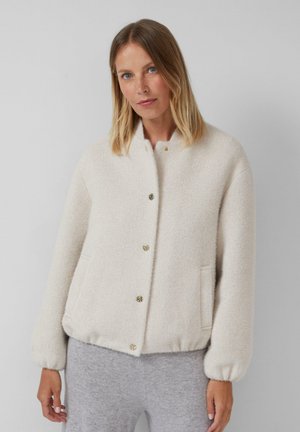 Veste texturée de couleur crème, avec un col rond, des boutons dorés et des poignets élastiques. Elle dispose de poches latérales et d'un design décontracté.