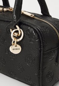 Crna torbica s teksturiranim dizajnom i zlatnim detaljima, utisnutim logotipom, dvostrukim ručkama na vrhu i odvojivim privjeskom koji prikazuje naziv brenda "GUESS."