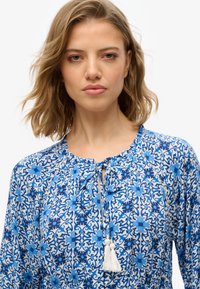 Niebieska bluzka w kwiatowy wzór z okrągłym dekoltem, sznurkiem do wiązania i detalami z frędzlami; lekką tkanina z misternymi wzorami.