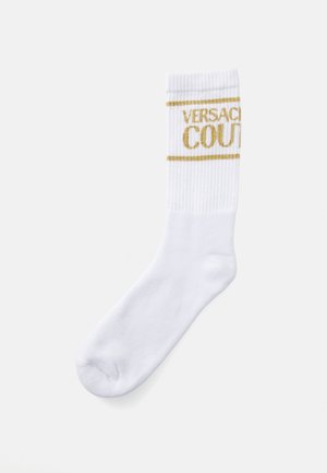 Calcetín blanco de estilo crew con puño acanalado y texto dorado "VERSACE COUTURE" y líneas cerca del borde superior.