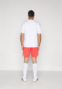 Biele športové tričko, koralové šortky, biele futbalové ponožky s čiernymi pruhmi a čierne kopačky s bielo-čiernym vzorom, pohľad zozadu.