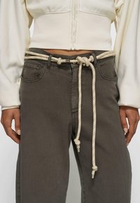 Baumwollseilgürtel um eine hoch taillierte dunkelgraue Hose mit Metallspitzen gebunden, getragen mit weißem, kurz geschnittenem Zip-up-Pullover.